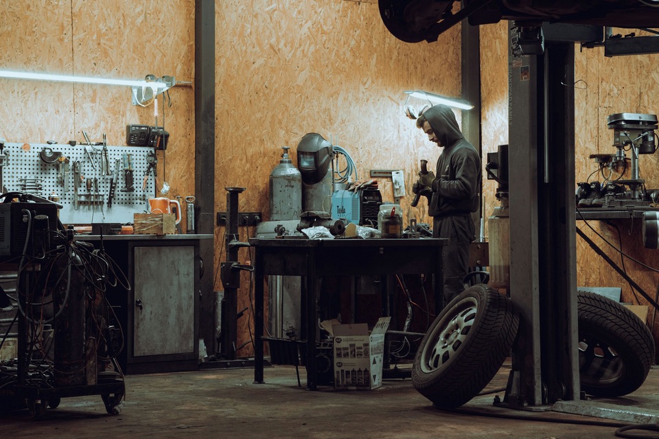 A mechanic working at a bench in a repair shop with a car lift, a tool wall and tires