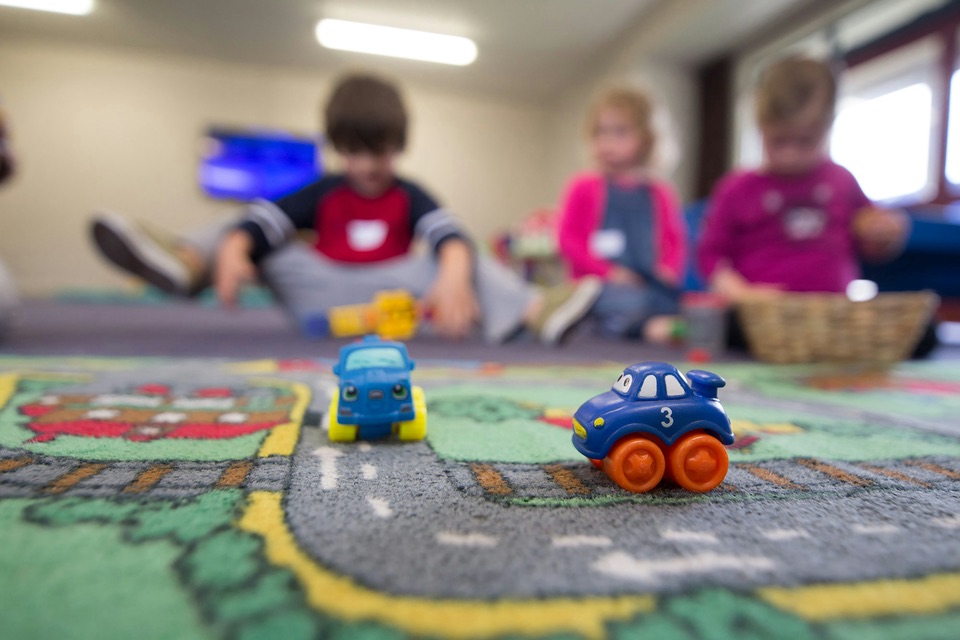 Toy cars on a play mat in a childcare room, with children playing out of focus behind