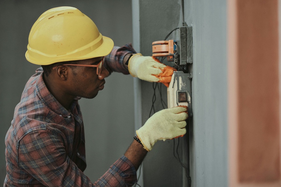 An electrician in a hard hat and gloves working on an electrical panel