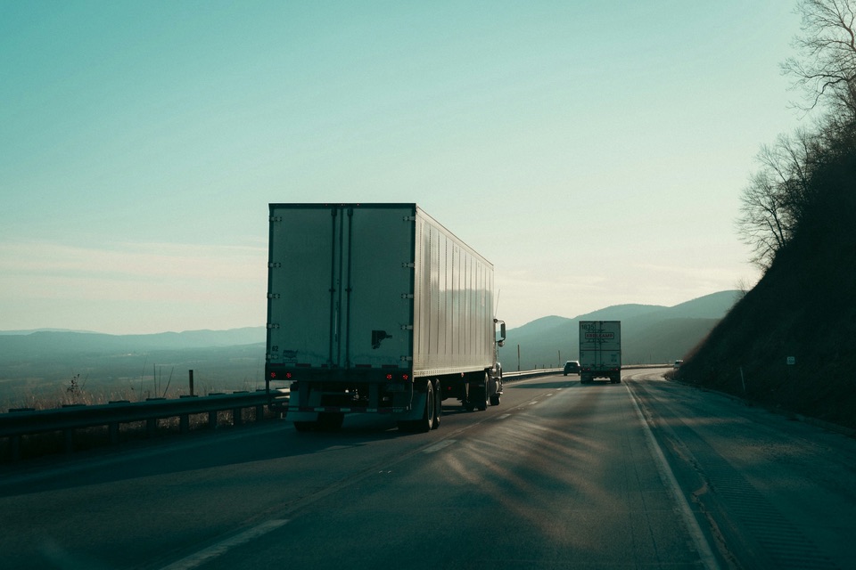 Two semi trucks with box trailers driving on a highway through hills
