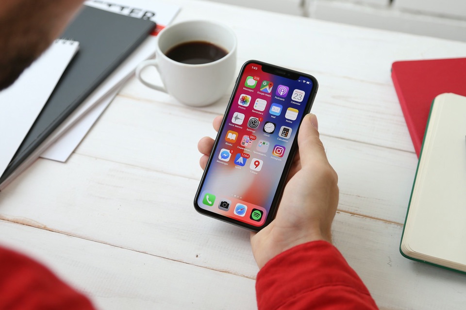 A hand holding an iPhone over a desk with a coffee cup and notebooks