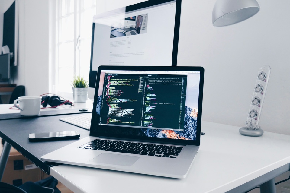 A laptop showing code on a bright desk beside a monitor
